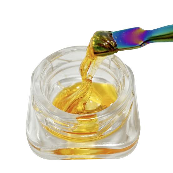 Golden translucent Delta 8 THC Shatter extract being pulled from a small clear glass container with a multi-colored metal dabbing tool