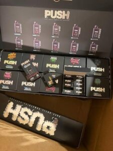 Box display container filled with authentic black push 2g disposable vape pens and colorful individual flavor boxes showing strain names like Peach Mango and Straw Guava.