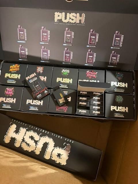 Box display container filled with authentic black push 2g disposable vape pens and colorful individual flavor boxes showing strain names like Peach Mango and Straw Guava.